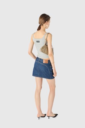 Back view of a woman in a gray sleeveless top and denim skirt, carrying a beige shoulder bag and wearing large hoop earrings; black heeled sandals visible at the feet.