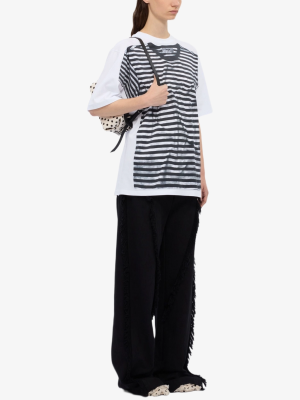 Model wearing a white oversized T-shirt layered with a black striped front panel, black wide-leg pants with fringe, and a polka-dotted bag over the shoulder.