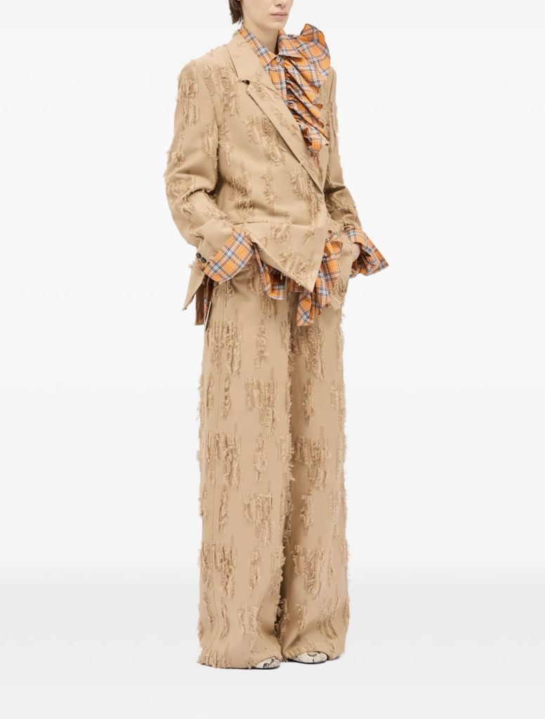 Model wearing a beige distressed blazer and matching wide-leg pants, with an orange plaid shirt peeking from the cuffs and neckline.