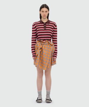 Model wearing a burgundy striped long-sleeve polo with an orange plaid wrap skirt, striped socks, and black-and-white sandals.