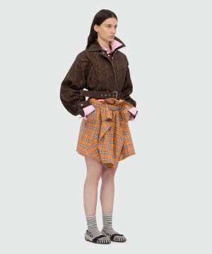 Model wearing a dark brown lace-patterned jacket with a pink shirt, paired with an orange plaid wrap skirt, striped socks, and black-and-white sandals.