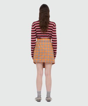 Person standing with back to camera, wearing a maroon and white striped long-sleeve top, orange plaid mini skirt, striped socks, and patterned sandals.