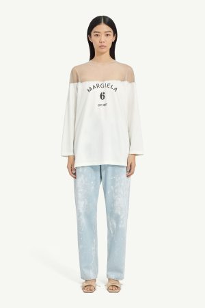 Model wearing a white off-shoulder top with sheer yoke and 'MARGIELA 6 EST 1997' print, paired with light blue splatter-jeans and beige sandals.