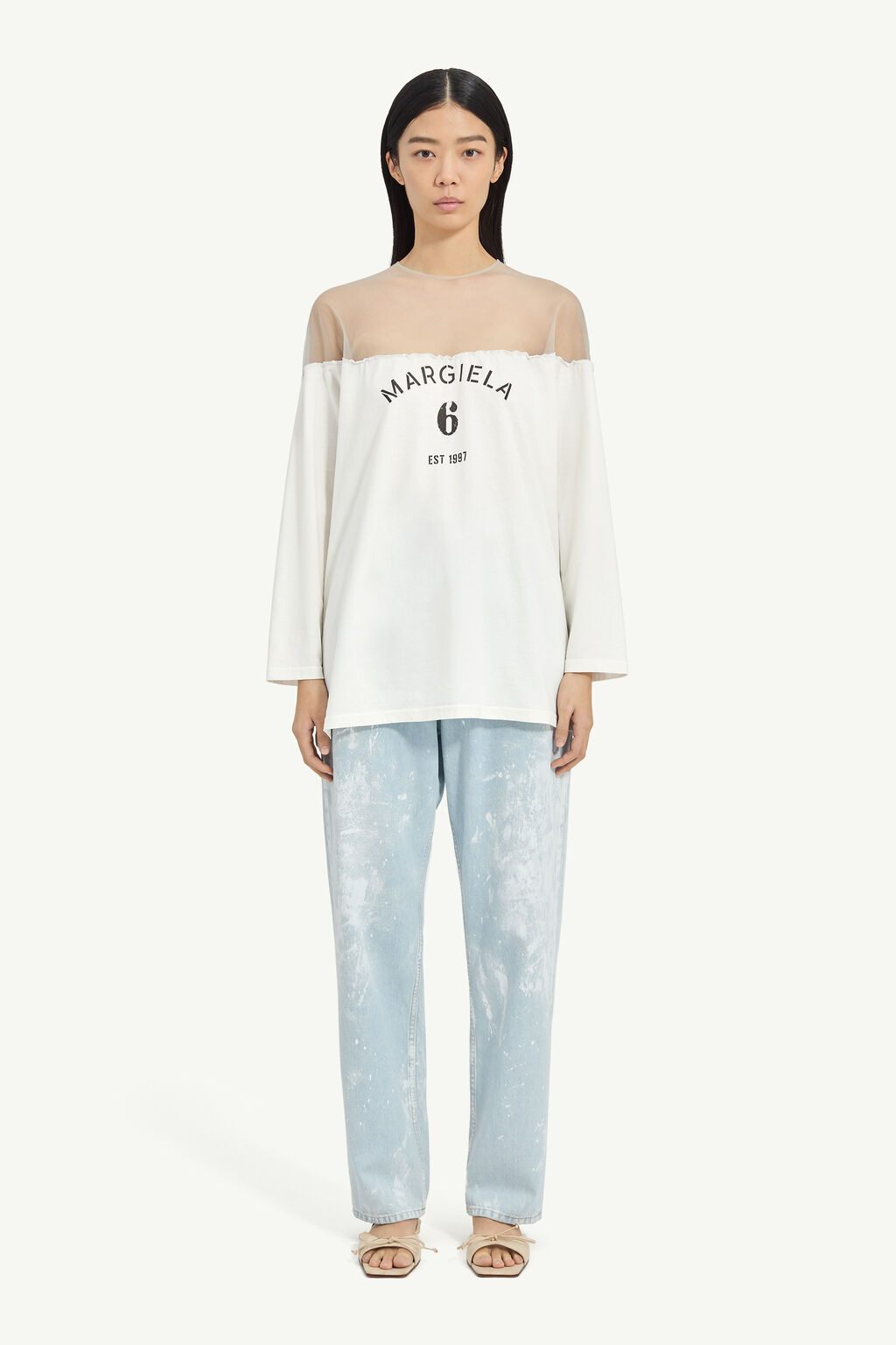 Model wearing a white off-shoulder top with sheer yoke and 'MARGIELA 6 EST 1997' print, paired with light blue splatter-jeans and beige sandals.