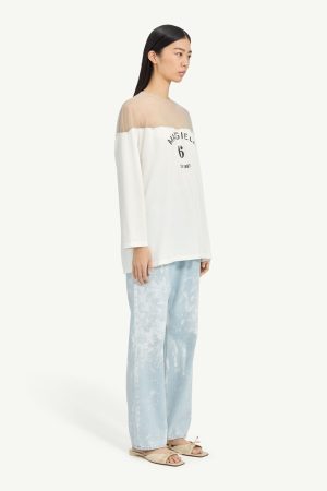 Side-view of a model wearing an off-shoulder white top with sheer yoke, black printed text, and light blue tie-dye jeans with beige sandals.