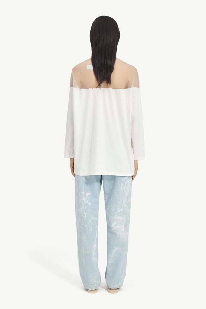 Back view of a person wearing an off-shoulder white top and light blue paint-splattered jeans, with long dark hair covering the shoulders.