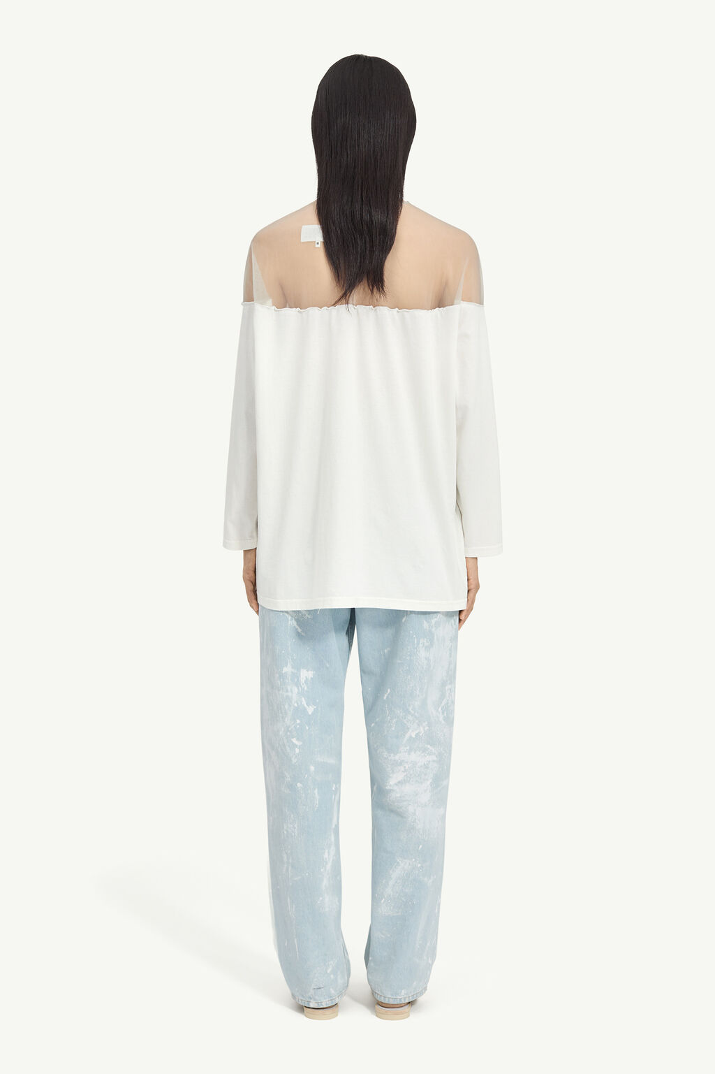 Back view of a person wearing an off-shoulder white top and light blue paint-splattered jeans, with long dark hair covering the shoulders.