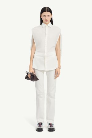 Model in an all-white ensemble: sleeveless oversized shirt, white pants, chunky sneakers with blue and pink laces, and a dark clutch in one hand.