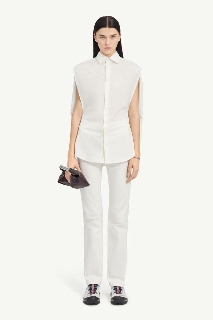 Model in an all-white ensemble: sleeveless oversized shirt, white pants, chunky sneakers with blue and pink laces, and a dark clutch in one hand.