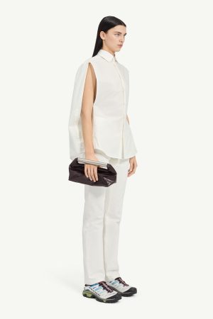 Model in a loose white sleeveless shirt and white pants, holding a black clutch with a metallic bar, wearing colorful athletic sneakers.
