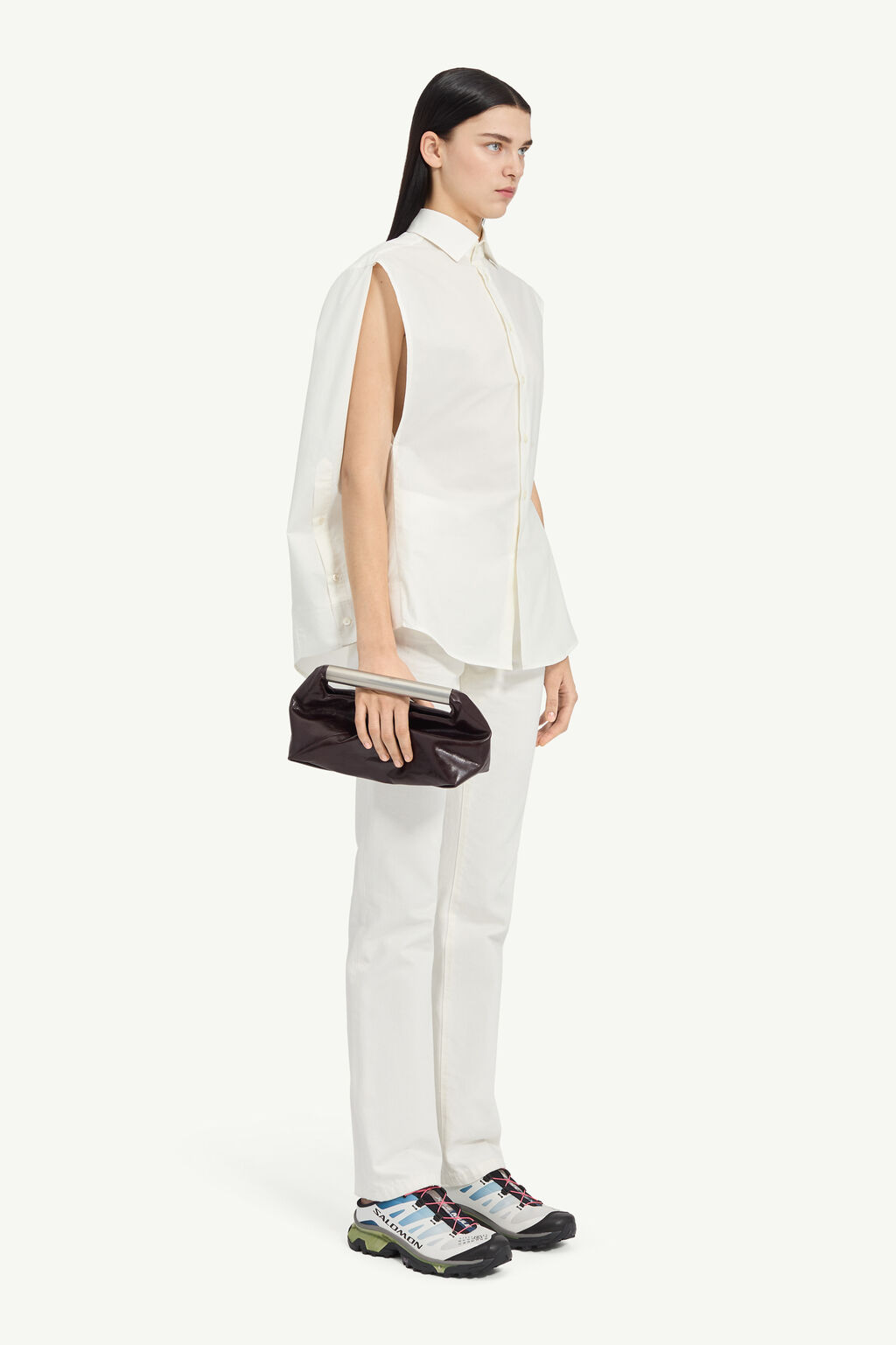 Model in a loose white sleeveless shirt and white pants, holding a black clutch with a metallic bar, wearing colorful athletic sneakers.