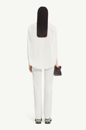 Person seen from behind in an all-white outfit: white shirt and pants, long black hair, holding a gray bag.
