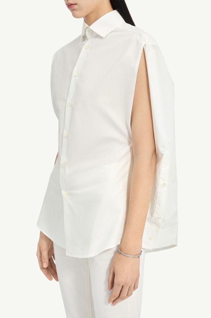 Side view of a white button-down shirt featuring a cape-like sleeve with an open shoulder slit, worn with white pants and a bracelet.