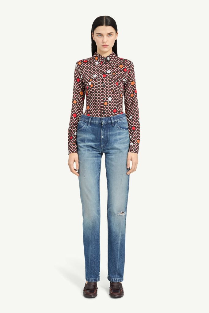 Full-body shot of a woman in a dark floral print button-down shirt tucked into blue distressed jeans, standing against a pale background.