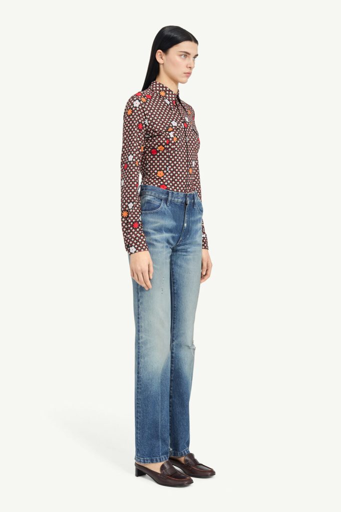 Profile view of a woman wearing a dark floral long-sleeve blouse, faded blue jeans, and brown loafers.