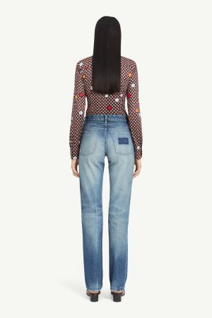 Back view of a woman with long black hair wearing a floral print sheer blouse tucked into light-wash straight-leg jeans, standing in heels