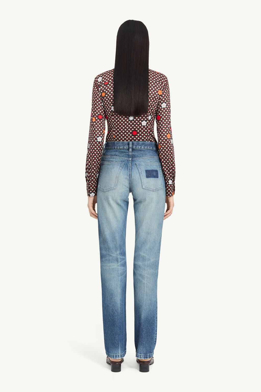 Back view of a woman with long black hair wearing a floral print sheer blouse tucked into light-wash straight-leg jeans, standing in heels