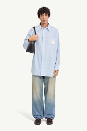Man wearing an oversized light blue striped shirt, wide-leg jeans, and brown loafers, carrying a small black handbag over the shoulder.