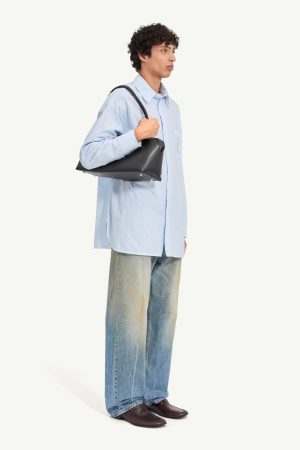 Young man in a light blue oversized shirt, bag slung over his shoulder, jeans and loafers, facing right side.