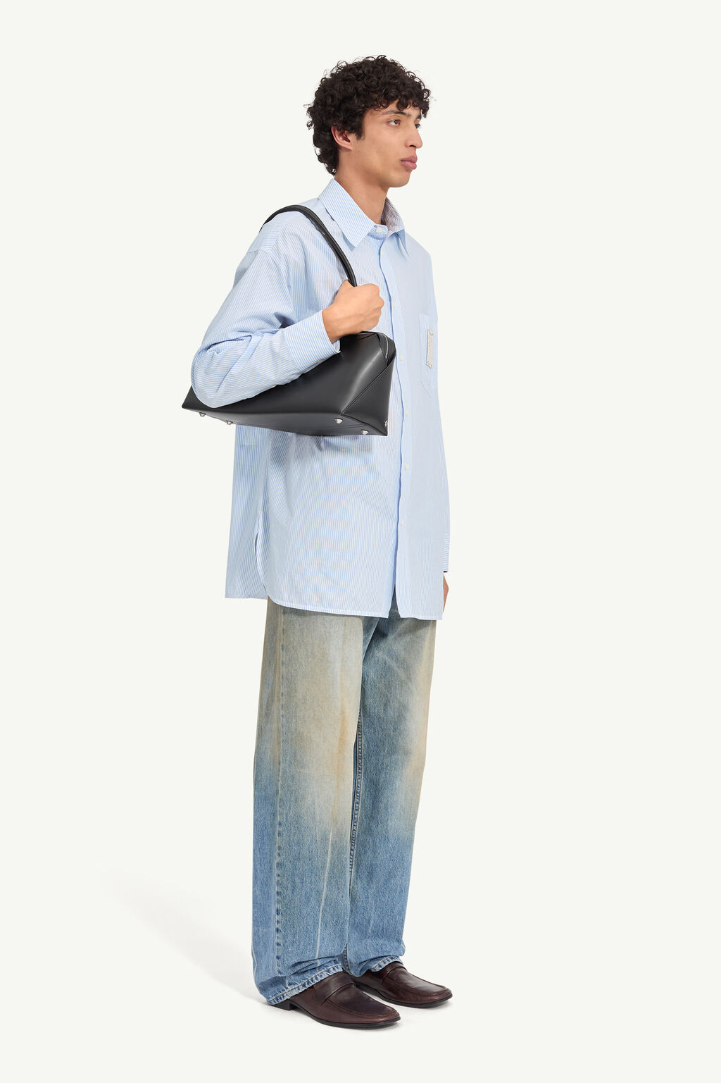 Young man in a light blue oversized shirt, bag slung over his shoulder, jeans and loafers, facing right side.