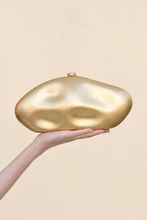 A hand holding a smooth, irregularly shaped gold sculpture against a pale beige background.