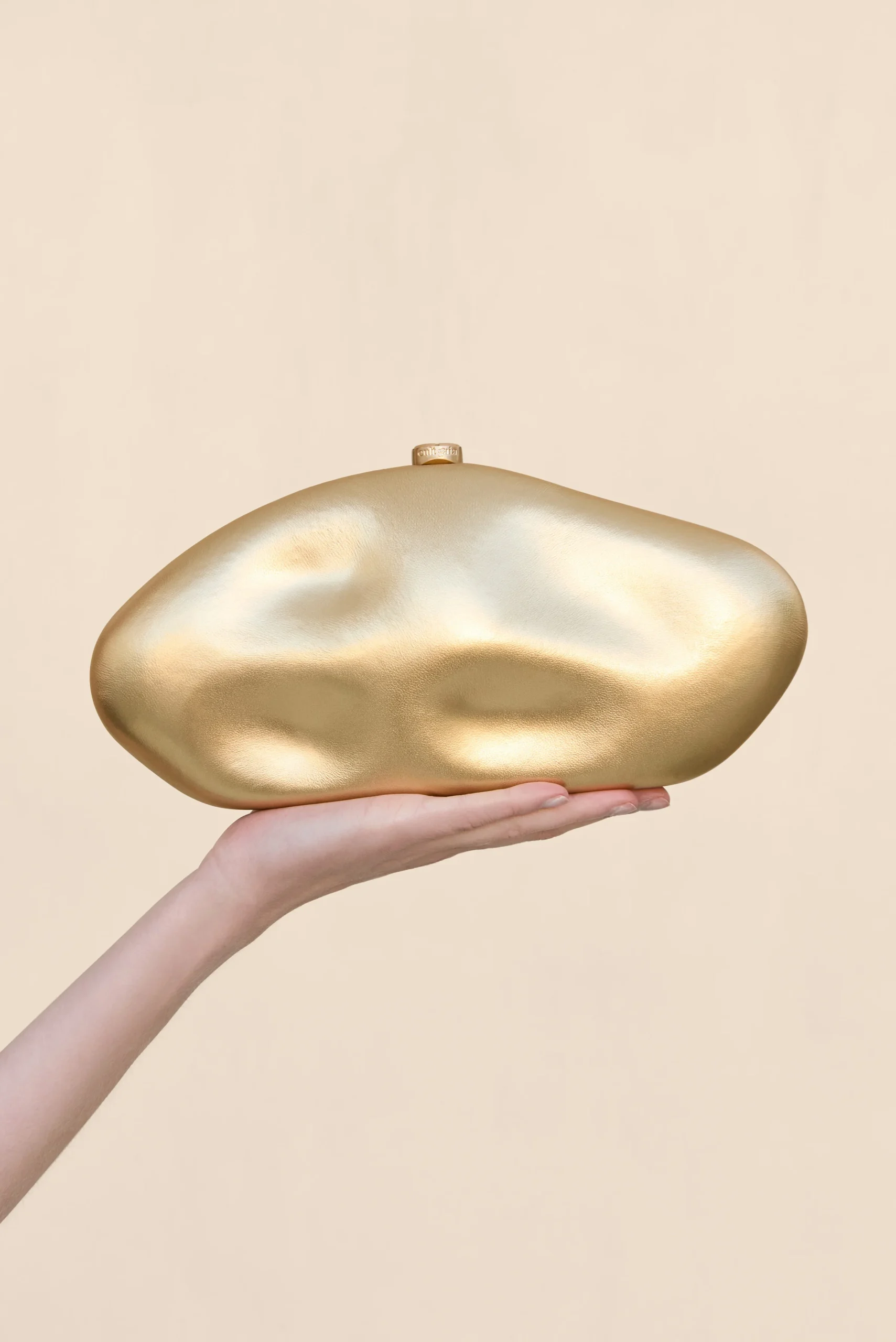 A hand holding a smooth, irregularly shaped gold sculpture against a pale beige background.