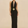 Model in a long black halter gown with a large gold circular belt buckle, standing against a beige backdrop.