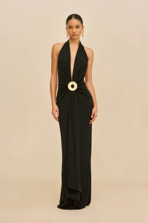 Model in a long black halter gown with a large gold circular belt buckle, standing against a beige backdrop.