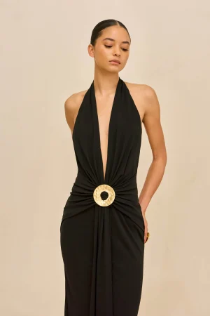 Woman in a black halter dress with a large circular gold belt, standing against a beige backdrop and looking downward.