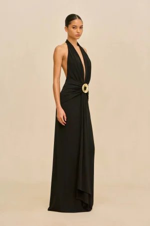 Full-length black halter gown with a deep V-neck and a gold circular belt buckle, standing against a beige backdrop.