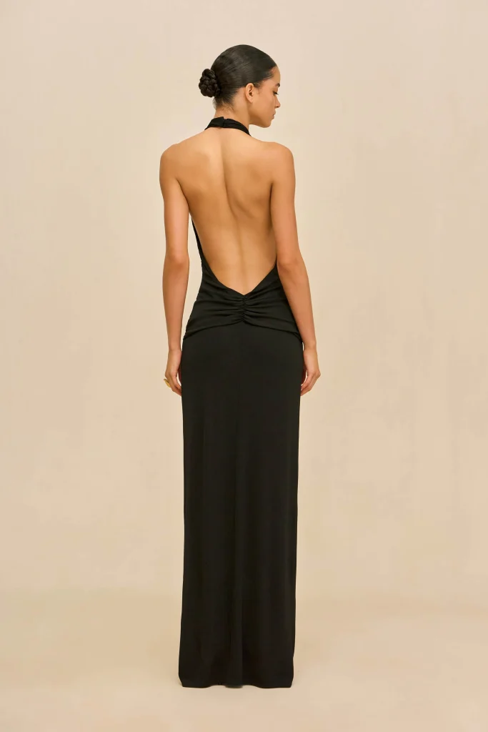 Back view of a woman in a black halter-back gown with a bun, standing against a beige backdrop.