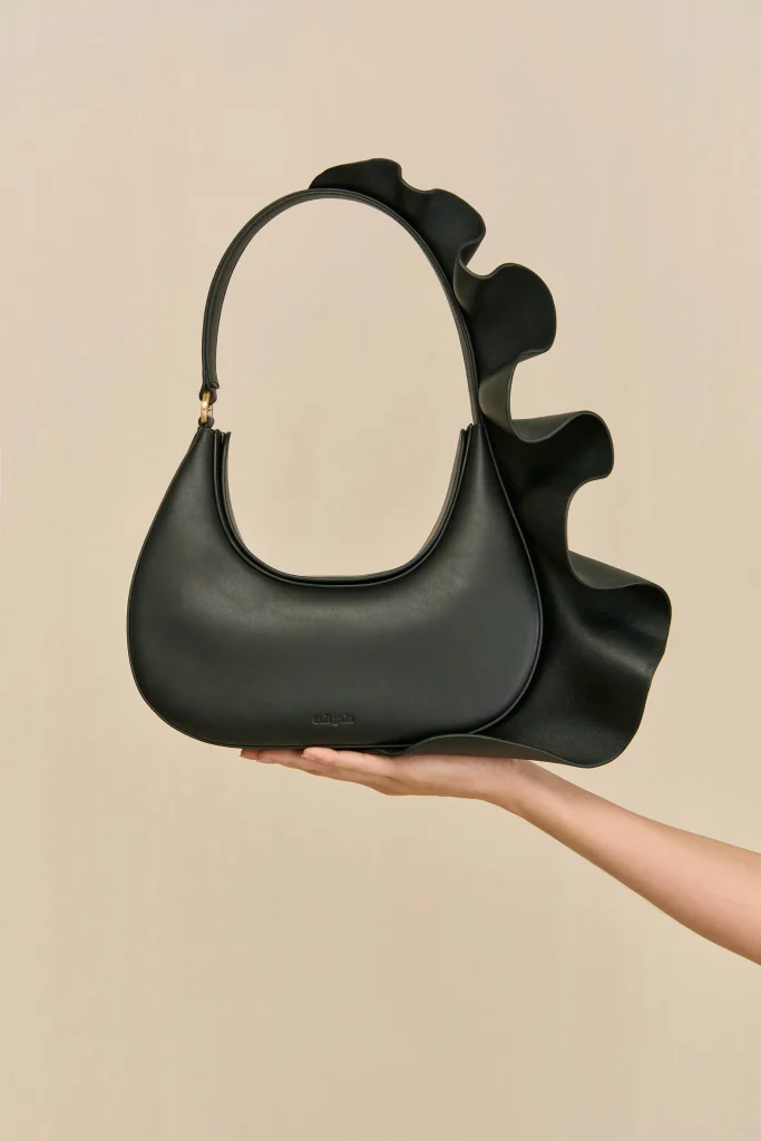 Black handbag with a large sculptural ruffle along the side, held by a hand.