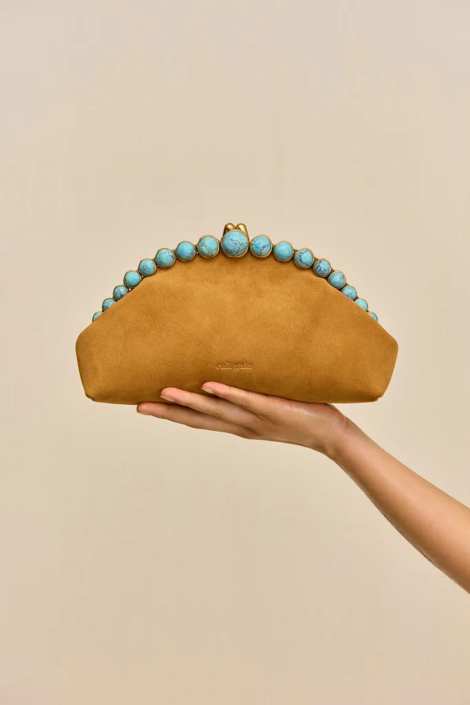 Tan suede clutch with turquoise bead trim held by hands against a beige background.