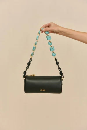 Hand holding a turquoise beaded strap above a black leather handbag against a beige background.