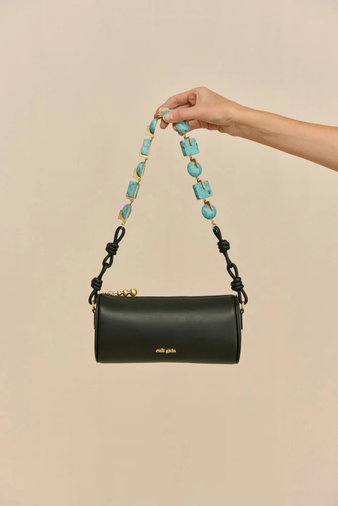 Hand holding a turquoise beaded strap above a black leather handbag against a beige background.