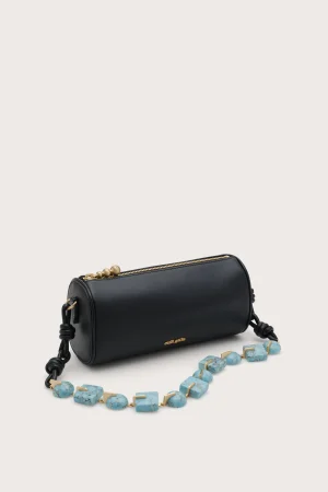 Black cylindrical handbag with a gold zipper, placed on a light background, with a turquoise bead necklace in front.
