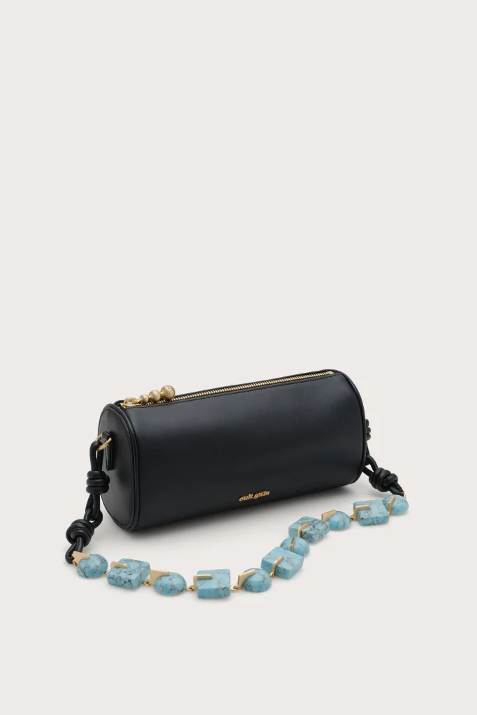 Black cylindrical handbag with a gold zipper, placed on a light background, with a turquoise bead necklace in front.