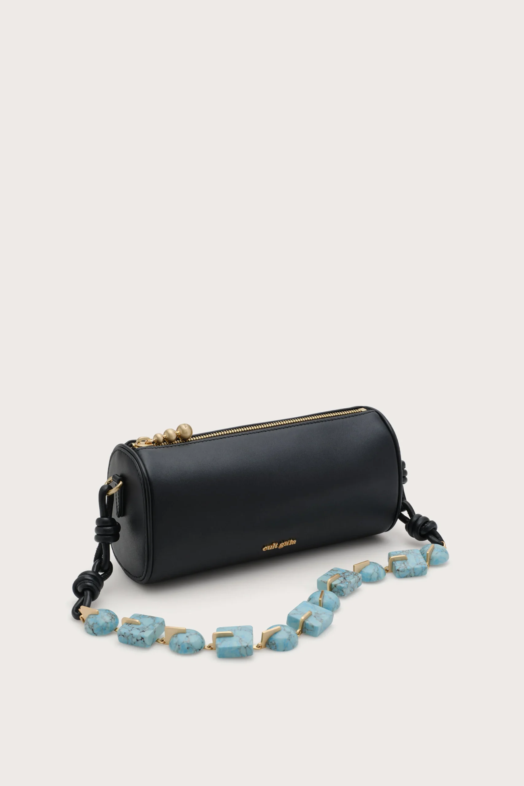 Black cylindrical handbag with a gold zipper, placed on a light background, with a turquoise bead necklace in front.