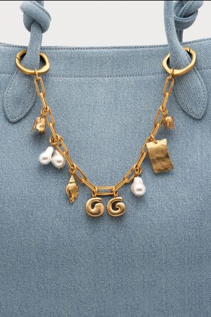 Close-up of a light denim bag with a gold chain necklace hanging from the handles, featuring pearls and charms including the letters GG.