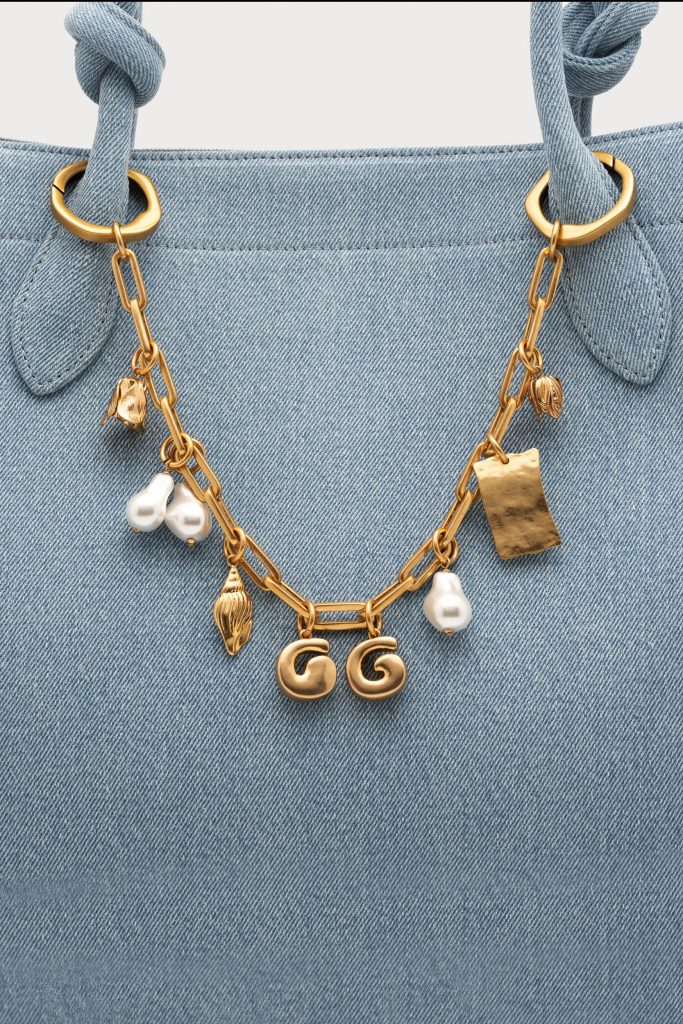 Close-up of a light denim bag with a gold chain necklace hanging from the handles, featuring pearls and charms including the letters GG.