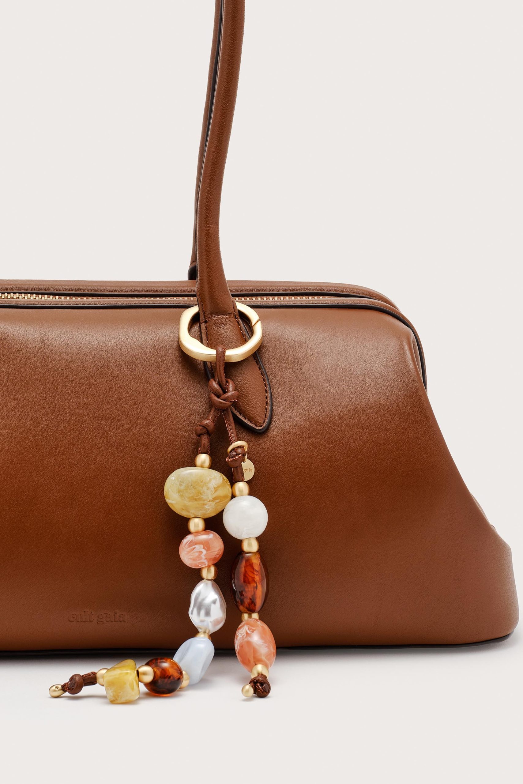 Close-up of a brown leather handbag with a gold ring hardware and a beaded accessory hanging from the strap surface edge.