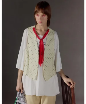 Model wearing a cream polka-dot vest over a white tunic with a red scarf, standing with one hand on a chair and holding a newspaper.