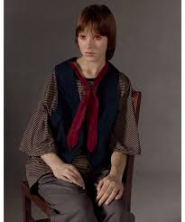 Person seated in a vintage-style outfit with a dark vest, red scarf, and checked sleeves against a gray background.