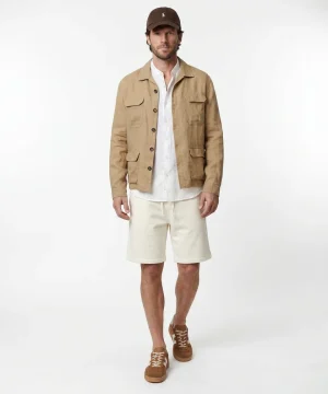 Male model wearing a tan utility jacket over a white shirt with ivory shorts, brown cap and matching sneakers on a studio backdrop.