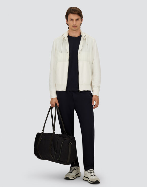 Man wearing a cream zip-up jacket over a navy T-shirt and navy trousers, holding a black duffel bag.