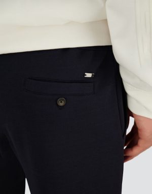 Close-up of dark navy trousers showing a back welt pocket with a single button and tiny metal logo above it.