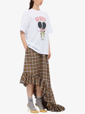 Model in a white oversized graphic T‑shirt with a broken-heart design, paired with a brown plaid high-low ruffled skirt, striped socks, and yellow heels.