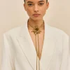 Close-up portrait of a woman with slicked-back hair wearing a white blazer and a gold sunburst necklace against a beige background.