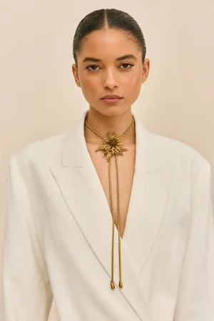 Close-up portrait of a woman with slicked-back hair wearing a white blazer and a gold sunburst necklace against a beige background.