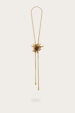 Gold necklace with a sunburst flower pendant hanging from a thin chain against a pale cream background.